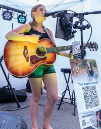 A young woman with blonde hair, wearing a black tank top and green denim shorts, performs live under a white tent canopy. She is playing an acoustic-electric sunburst guitar and singing into a microphone. To her right, on a stand, is an iPad and a tip sign with a photo of the performer, the text "Laura Rullo," "Tips Appreciated!", a QR code, and social media handles. Professional stage lighting trusses are visible behind her.