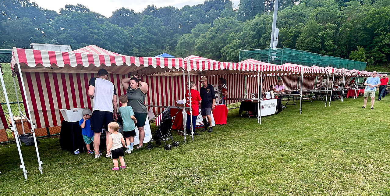 A wide-angle photograph captures a long row of identical red-and-white striped canopy tents set up in a grassy park. Several families and individuals are walking along the front of the tents, interacting with people at the booths. On the far left, a man in a white tank top and black shorts is standing with his back to the camera, talking to a woman holding a baby. Two small children are nearby. A black stroller is parked next to the booth. Further down the line, other vendors are visible. Behind the tents, there is a chain-link fence and a dense forest of green trees. The sky is overcast.