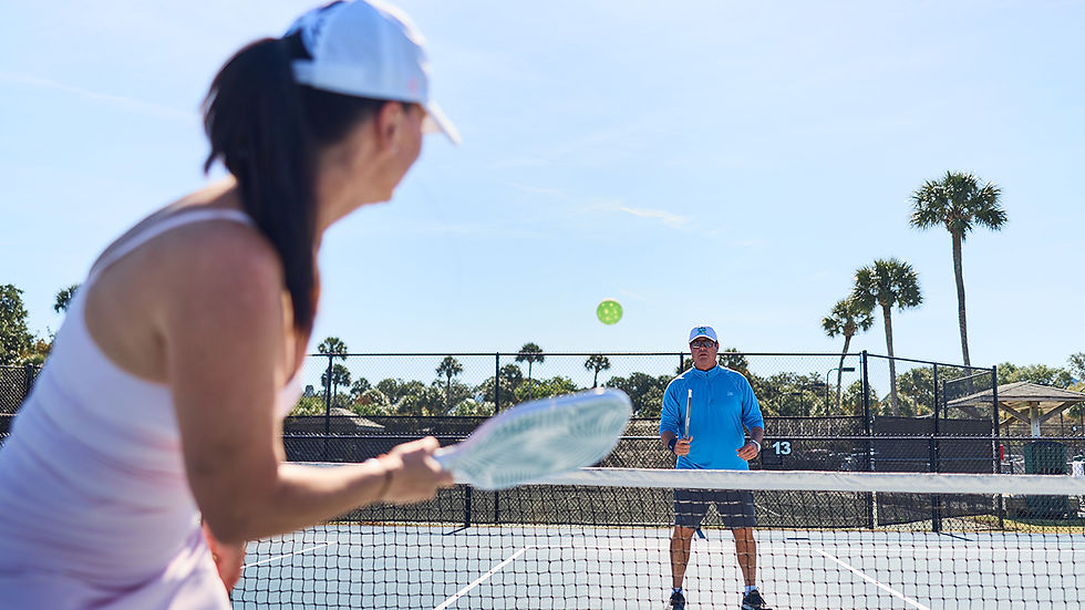 Two adults playing pickleball on an outdoor court at Wild Dunes Resort, with palm trees and blue sky in the background. A woman in the foreground is swinging a white paddle toward the net, and a man in a blue shirt is ready to receive the shot.
