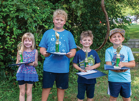 Four children, three boys and one girl, stand side-by-side in front of a leafy green hedge. They are each holding a trophy with a fish on top and a certificate. The girl on the far left, with a big smile, wears a blue patterned top and shorts. The boy next to her, who is taller than the others, wears a blue t-shirt and dark shorts. To his right, a boy in a dark blue t-shirt with a monster graphic smiles at the camera. The boy on the far right wears a light blue t-shirt, shorts, and a colorful baseball cap.