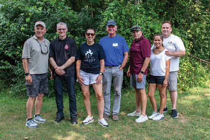 A group of six adults stands in a line facing the camera, with a dense green forest behind them. From left to right: a man in a tan shirt and cargo shorts; a man in a black polo shirt; a woman in a black t-shirt and white shorts; a man in a blue polo shirt; a man in a dark red polo shirt and gray shorts; and a woman in a white t-shirt and dark shorts standing next to a man in a white t-shirt.