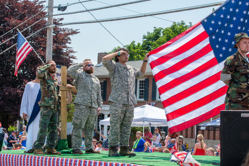 A group of men in military camouflage stand on a parade float and salute as a large American flag waves behind them.