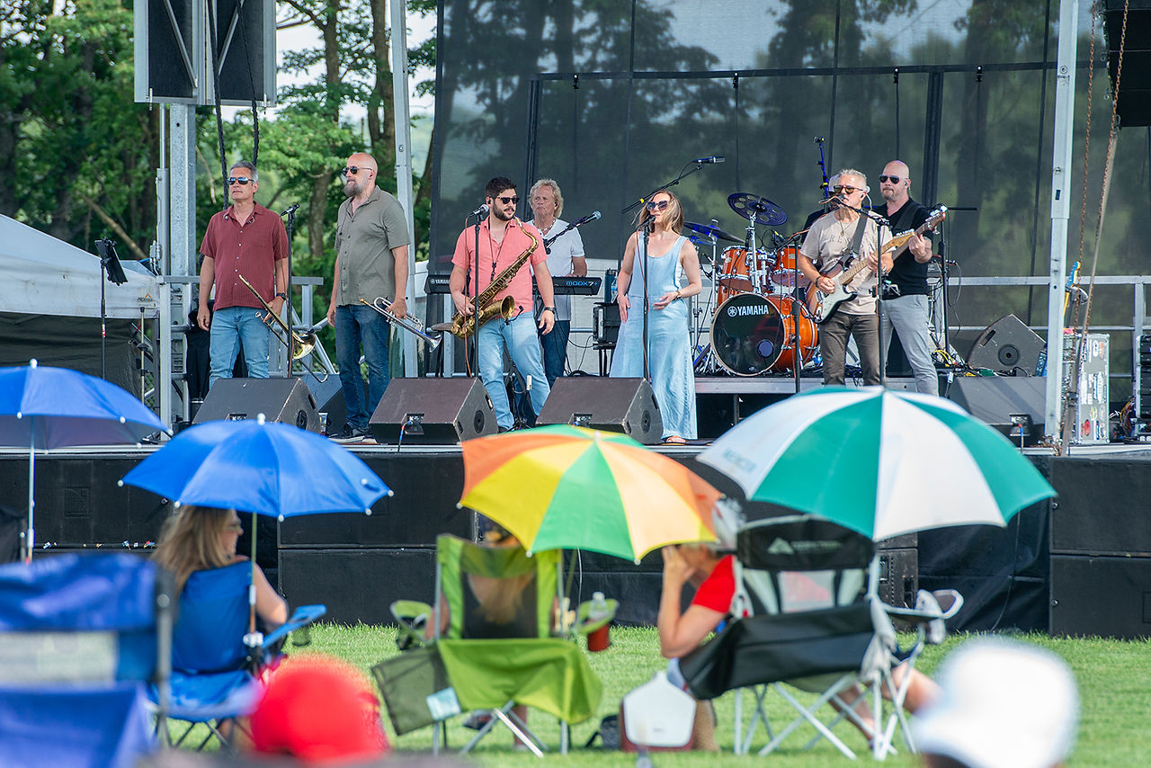 A band performs on an outdoor stage at a festival, with people watching from chairs under umbrellas in the foreground.