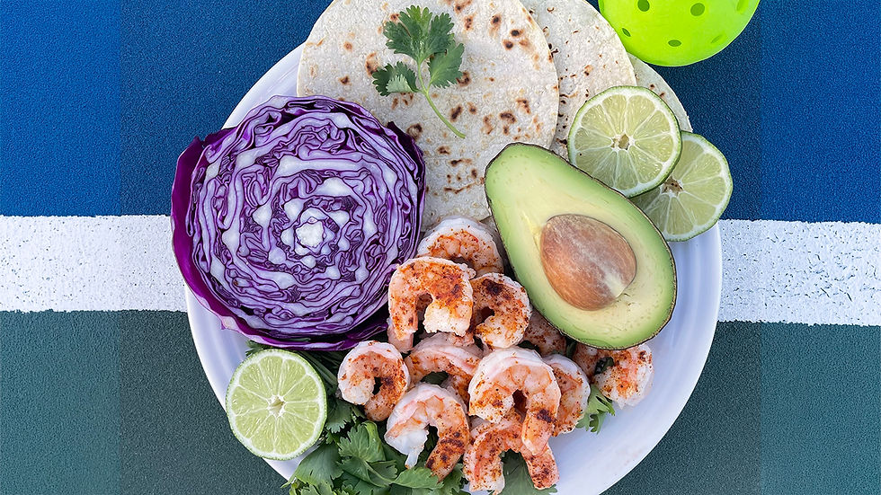 Overhead view of a white plate featuring seasoned shrimp, corn tortillas, a halved avocado, sliced red cabbage, lime wheels, fresh cilantro, and a bright green pickleball on a blue and green court background.