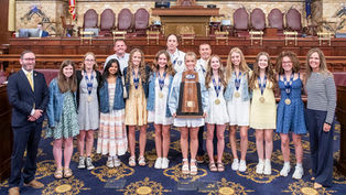 A photograph shows the Norwin Girls Cross Country Team standing together inside a government chamber. Ten young women stand in a row wearing dresses, with most also wearing medals around their necks and denim jackets. They are all smiling at the camera, and the girl in the center holds up a large wooden PIAA championship plaque. To the far left stands a man in a dark suit and yellow tie, and to the far right stands a woman in a striped long-sleeve shirt and dark trousers. Three coaches stand directly behind the team. The group is posed on a patterned blue carpet in front of rows of historic dark wood desks and elevated leather chairs.