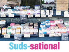 A detailed outdoor market display for Pittsburgh Artisanal Soap Co. featuring rows of colorful, handmade soap bars arranged on tiered shelving. The table is draped in a blue cloth, with clear signage indicating various scents like "Ocean," "Fruity," and "Floral," alongside a white banner that reads, "Experience the Beauty of Soap!"