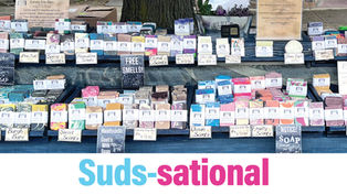 A detailed outdoor market display for Pittsburgh Artisanal Soap Co. featuring rows of colorful, handmade soap bars arranged on tiered shelving. The table is draped in a blue cloth, with clear signage indicating various scents like "Ocean," "Fruity," and "Floral," alongside a white banner that reads, "Experience the Beauty of Soap!"