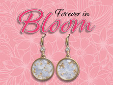 Two round, gold-rimmed drop earrings featuring small, light blue pressed flowers and gold flakes encased in clear resin, set against a pink floral-patterned background with the text 'Forever in Bloom' above them.
