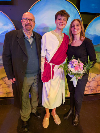 A family portrait on stage at Carnegie Stage featuring the creative team behind "Another Miracle." On the left is composer Charles "Chuck" Sperry in a long dark coat and glasses. In the center is his grandson, Jude Glover, in his white and red costume as Jesus. On the right is his daughter, Nicole Glover, wearing a black top and holding a bouquet of pink and purple flowers. They stand together in front of the painted blue arched backdrops, symbolizing the family’s three-generation collaboration.