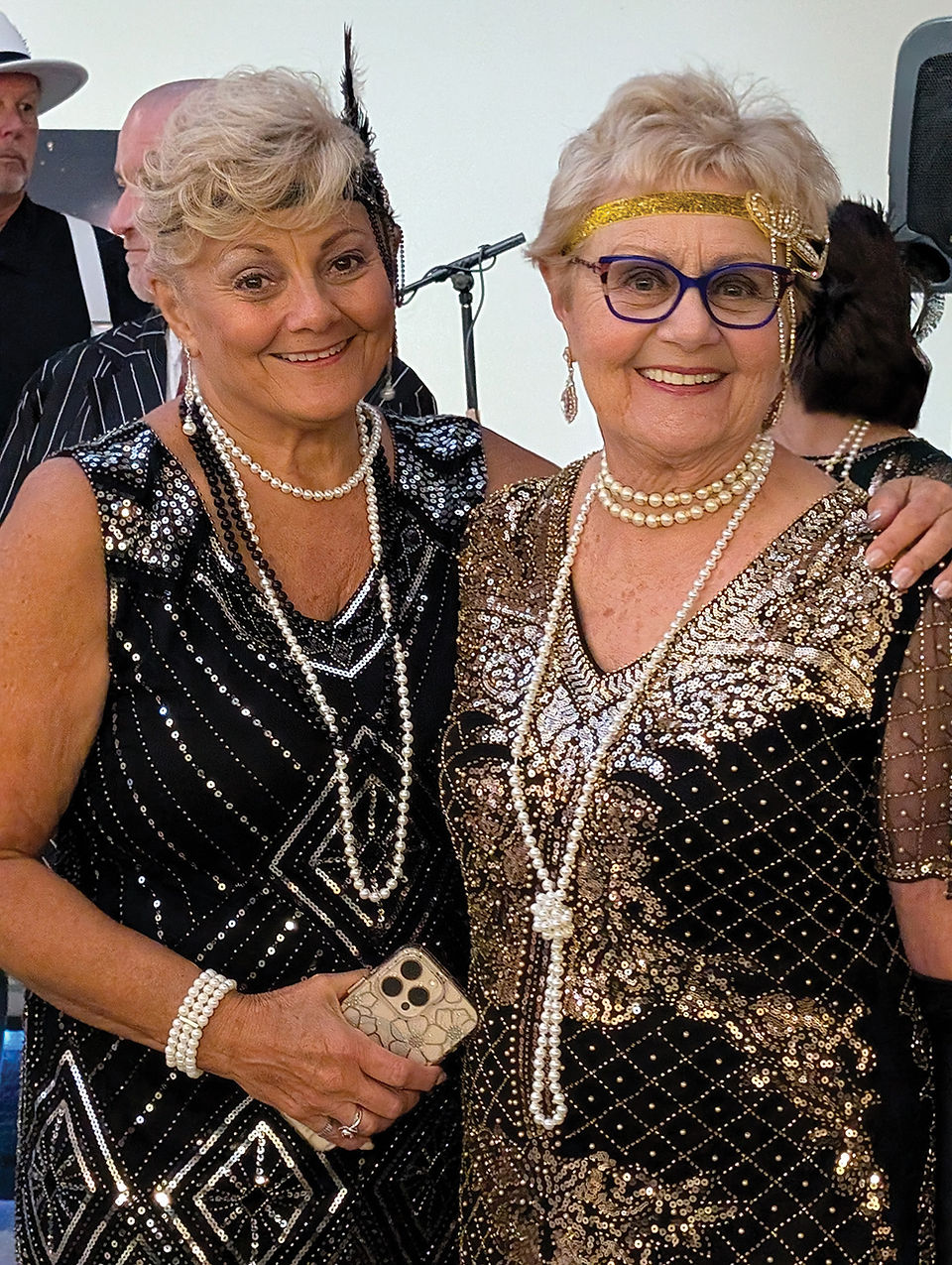 Two women smiling closely together, both wearing beaded flapper dresses and multi-strand pearl necklaces.