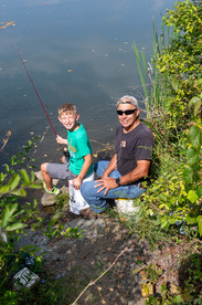 A young boy with light hair and a green shirt sits on a white bucket on the bank of a pond, holding a fishing pole. An older man sits on a bucket beside him, smiling.