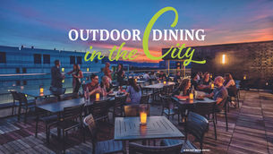 A rooftop patio scene at Eliza Hot Metal Bistro during twilight, featuring guests seated at metal tables illuminated by small candles. A vibrant sunset glows over the city skyline in the background. A large, stylized text overlay reads: "OUTDOOR DINING in the City."