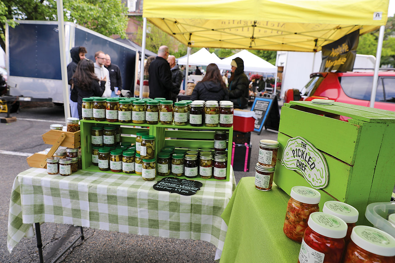 A market stall table draped in a green-and-white checkered cloth is packed with numerous jars of pickles and other pickled vegetables from "The Pickled Chef." The jars are displayed on and within green wooden crates. A chalkboard sign in front of the pickles reads "Local, Small Batch, Scratch Made, Pickles + Veggies, 16oz Pint - $9 Jar." To the right, a large green crate bears the brand's logo, and several jars of kimchi are visible in the foreground. In the background, people mingle under yellow and white vendor canopies.