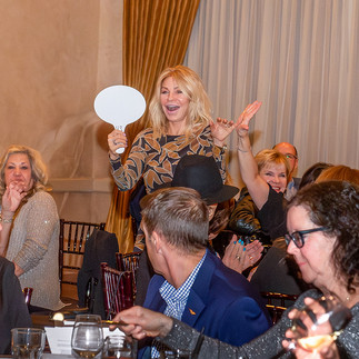  Gemini said A woman in a leaf-patterned dress smiles excitedly while holding up an auction paddle among a crowd of seated guests during the live auction portion of the 17th Annual Symphony of Food.