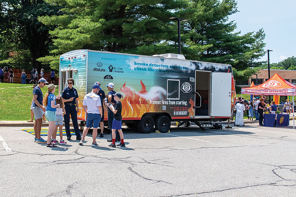 A crowd of people, including firefighters, gather around a large, colorful trailer labeled with fire safety information and sponsors like Allstate and St. Clair Hospital.