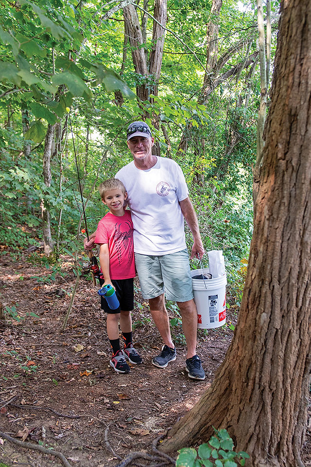 A man with a baseball cap and a boy in a red shirt stand next to a tree on a wooded path. The man holds a fishing pole and a white bucket, and the boy holds a blue cup and a fishing pole.