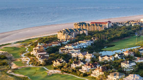 An aerial view of the Wild Dunes Resort showing a large beachfront hotel, numerous multi-story luxury homes and villas, and parts of a manicured green golf course bordering the wide, sandy beach and the blue ocean.