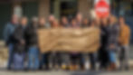 A diverse group of adults stand outdoors in winter clothing, posing with a large, hand-drawn paper sign. The sign, which is held up across the center, reads in black ink: 'ROOTS TO HOME AFFORDABLE HOUSING PROJECT' with two small heart symbols. The group consists of about fifteen people of various ages and races, including: a man with a knit hat on the left, a man in a black coat to his left, and a man in a long, dark coat. A woman with glasses and a scarf is central, and on the right is a tall man in an orange-brown jacket and a woman in a long black puffer coat. Behind them is a building facade, and to the right, a prominent 'DO NOT ENTER' sign is visible. The ground is dry asphalt with some snow patches.