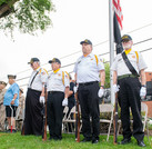 A medium shot, taken from a low angle, of a four-man veterans' honor guard standing at attention in a grassy area. The men wear white uniform shirts with short sleeves, black trousers, and various hats: three wear dark blue caps with yellow insignia, and the fourth (further back on the left) wears a tan garrison cap with a "VFW" logo. They wear black belts and some have gold lanyards and white gloves. One veteran on the right is at attention with a rifle butt on the grass; another on the left holds a walking cane. A flagpole with the American and a black POW/MIA flag is visible to the right. The background shows tree leaves, utility lines, and the corner of a brick building.