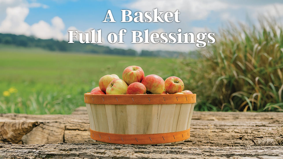 A rustic, light-wood stave basket with orange-painted rims, filled with red-and-yellow-streaked apples, sits on a rough-hewn log. The blurred background shows a sunny, green pasture under a partly cloudy sky. The white, stylized text "A Basket Full of Blessings" is centered over the top of the image.