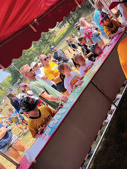 Children and adults play a carnival game at a red-and-white striped tent. A boy in a Steelers jersey is in the foreground, focused on the game.