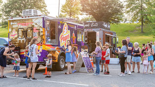A vibrant, late afternoon photograph capturing the bustling atmosphere of the 2026 Peters Township Market in the Park. The scene is set in an asphalt parking lot, with a verdant, tree-covered hillside rising behind the various vendor trucks. The central focus is a colorful food truck called 'Page's Pure Ice Cream', a retro-style van with a predominantly purple exterior and yellow and white accents. Large illustrations of ice cream cones and the name 'Page's' are painted on the side, along with smaller vintage photographs and text, including 'Making Pittsburgh Sweeter Since 1951'. Next to Page's is another vendor stall, 'Good Eats', in a more rustic, corrugated metal trailer. A line of customers, a mix of adults and children, snakes in front of the trucks. In the foreground, a woman in a navy blue dress helps a young girl in a patterned dress and hat manage a cup. A woman in a tie-dye tank top stands further along, followed by other families. An A-frame menu board stands in front of Pa