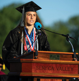A medium shot of a female graduate with long straight hair delivering a speech from a wooden podium featuring the "SENECA VALLEY" inscription. She is wearing a black cap and gown alongside white honor cords and a thick red, white, and blue medal ribbon around her neck. She speaks into a microphone with a blue foam cover under clear daylight.