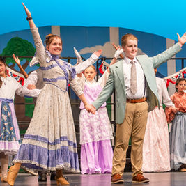 A medium shot of a large group of young performers on stage, striking a dynamic pose. A girl in a patterned dress and a boy in a light green suit are in the center, holding hands and extending their other arms wide. Other cast members flank them, cheering with gloved hands raised. The background includes stylized blue and white striped flats.