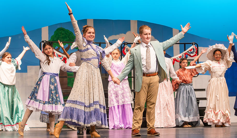 A medium shot of a large group of young performers on stage, striking a dynamic pose. A girl in a patterned dress and a boy in a light green suit are in the center, holding hands and extending their other arms wide. Other cast members flank them, cheering with gloved hands raised. The background includes stylized blue and white striped flats.