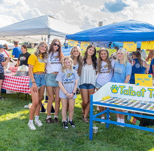A group of seven girls, mostly teenagers, smiles while posing for a photo in front of a colorful painted wooden cutout board.
