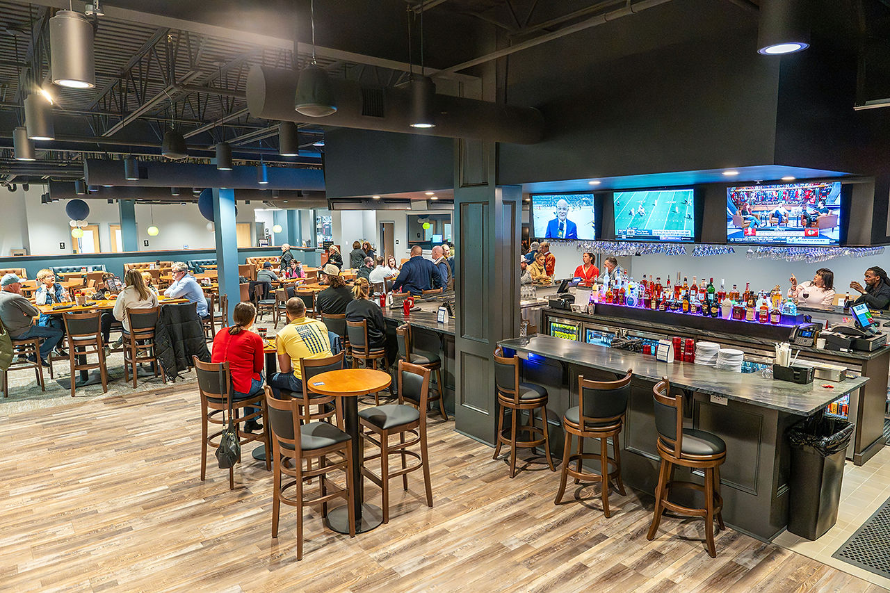 A lively view of the bar area at Baseline Bar & Grille. Patrons sit at a sleek, dark L-shaped bar with high-back stools. Three large television screens above the bar display sports and news programs. In the background, additional guests are seated at dining tables in a spacious room with wood-look flooring and modern industrial lighting.