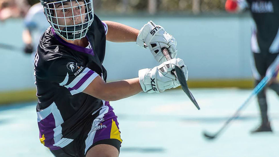 A close-up shot of a player playing field hockey with protective gear on. The background features other players on a blue surface. They wear a black helmet with a white mask, a white mask, black and purple shorts, black and white socks, and white gloves, holding a field hockey stick in both hands with their right arm extended.