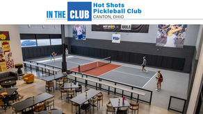 An indoor pickleball court with two players on it. The facility has high ceilings, a lounge area with tables and chairs, and large banners on the walls with images of pickleball players.
