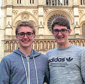 A sunny, outdoor portrait of two young men, presumably brothers, standing in front of the ornate, stone façade of Notre Dame Cathedral in Paris. Both wear glasses and smile for the camera. The man on the left wears a grey hoodie, and the man on the right wears a grey sweatshirt with a black "adidas" logo.
