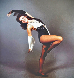 An archival photo of a young Adrianne Arena-Jacobs performing a jazz dance pose, wearing a black and white sequined costume and fishnet stockings, early in her dance career.