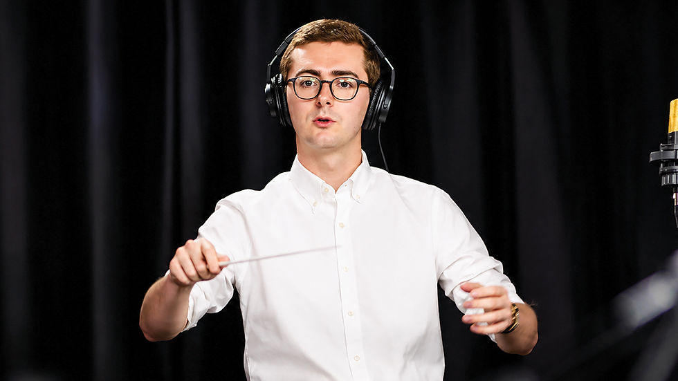 A medium shot of a young man standing against a plain black curtain backdrop. He has light brown hair, wears wire-rimmed glasses and large black headphones, and is dressed in a white button-down shirt. He is actively conducting with a baton in his right hand, looking directly at the camera with a neutral expression. A professional-grade microphone is visible on a stand to the right.