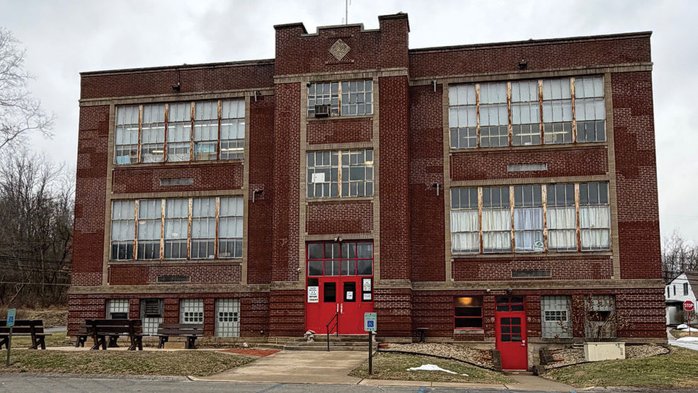 A wide, straight-on exterior shot of the historic Venetia School, a three-story red brick building with a symmetrical design and large multi-paned windows. The central entrance features bright red double doors under a crenelated brick parapet. A small paved walkway leads to the entrance, and several dark wooden benches are situated on the lawn to the left. The sky is overcast, and the surrounding landscape shows bare trees and patches of melting snow.