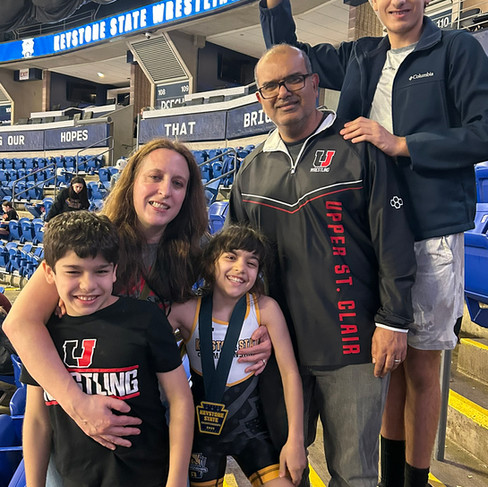 A smiling Maya Bhandari posing with her family in the stands at the Keystone State Wrestling Championships. She is wearing her wrestling singlet and a championship medal, surrounded by her parents and two brothers, with the arena seating and event banners visible in the background.