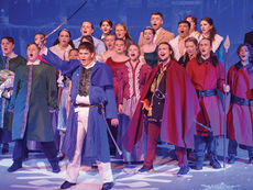 A dramatic, full-ensemble stage shot featuring the citizens and guards of Arendelle. In the front center, a teenage actor portraying Hans stands in a commanding pose, wearing white trousers and a blue long-coat with a capelet, holding a silver sword high in his right hand. To his right, the Duke of Weselton stands in a red cape and black jacket, also singing. On the far right, two guards in long red tunics hold silver-tipped spears. Behind them, a large group of cast members in various townsperson costumes—including dresses, waistcoats, and cloaks—all have their mouths open in song with intense expressions. The stage floor is decorated with a large white snowflake decal.
