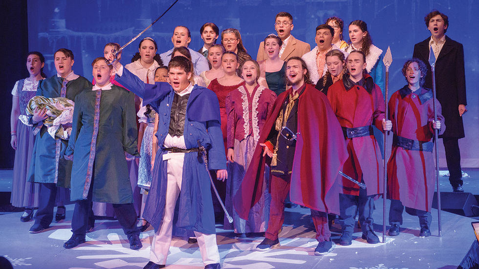 A dramatic, full-ensemble stage shot featuring the citizens and guards of Arendelle. In the front center, a teenage actor portraying Hans stands in a commanding pose, wearing white trousers and a blue long-coat with a capelet, holding a silver sword high in his right hand. To his right, the Duke of Weselton stands in a red cape and black jacket, also singing. On the far right, two guards in long red tunics hold silver-tipped spears. Behind them, a large group of cast members in various townsperson costumes—including dresses, waistcoats, and cloaks—all have their mouths open in song with intense expressions. The stage floor is decorated with a large white snowflake decal.