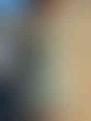 A young woman posing in a sage green dress designed by Sidney Rice. The dress has a textured lace bodice with corset-style boning and sheer tie-shoulder straps. She wears a white floral corsage and stands outdoors near a river with Pittsburgh's iconic yellow bridges and city buildings in the background.