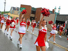 A street-level photograph capturing a line of seven young female cheerleaders marching in a parade on a wet, overcast day in a town center. The girls wear matching red and white sleeveless uniforms with "North Hills" across the chest, white skirts, and white sneakers. They hold red and white pom-poms, some raised in the air. The leading cheerleader smiles directly at the camera. In the background are brick buildings, a "Welcome to..." sign, old-fashioned street lamps, a few spectators, and a speed limit sign.