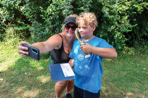 A woman with blonde hair wearing sunglasses and a black tank top kneels next to a young boy with curly blonde hair. They are taking a selfie with her phone. The boy holds a golden trophy and a blue folder with a certificate. They are in a grassy area with a dense green hedge behind them.