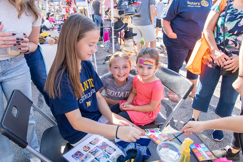 A young woman sitting on a folding chair painting a rainbow face design on the cheek of a small girl in a coral-colored dress. The face painting station is part of the festival activities.