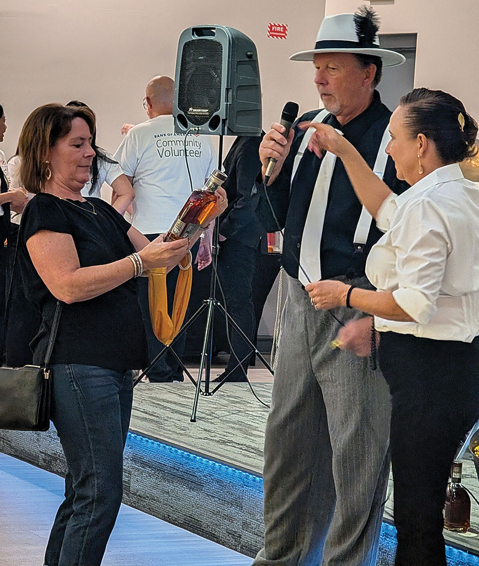 A guest holds up a bottle of liquor while interacting with a man and woman dressed in 1920s attire.
