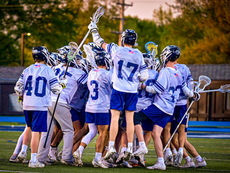 A group of McDowell Trojan lacrosse players in white jerseys with blue numbers huddle together in a celebratory circle on the field at sunset. Player number 17 is seen from the back, jumping slightly with his lacrosse stick raised high in the air, capturing the high-energy "fastest game on two feet" atmosphere described in the article.
