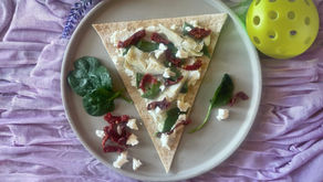 A top-down photograph captures a single, triangular wedge of a flatbread pizza, presented on a textured, light-gray round plate. The base is a whole-wheat flatbread, generously topped with creamy white feta cheese crumbles, dark-red sun-dried tomato slivers, and tender, light-green artichoke heart quarters. Several fresh, vibrant green spinach leaves are nestled within the toppings on the flatbread, with a few additional loose leaves, a small pile of crumbled feta, and a couple of sun-dried tomato pieces arranged artfully on the plate below the pizza wedge. The plate rests on a heavily folded, light purple, slightly crinkled textile surface. In the top-left corner, a few sprigs of purple lavender flowers are visible. A bright, perforated yellow pickleball is partially visible in the top-right corner, near the edge of the plate. The natural light illuminates the scene from the left, highlighting the textures of the food and fabrics. The composition is centered and overhead. The entire a
