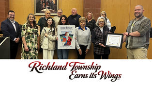 A medium wide shot in color shows 11 people in casual clothing smiling in front of wood paneled walls in a meeting room, holding a framed certificate and a rectangular sign with a Keystone shaped logo in the center and the words "BIRD TOWN PENNSYLVANIA birdtownpa.org" below the logo. At the top of the sign in large black capital letters are the words "BIRD TOWN PENNSYLVANIA". In the center of the sign is a blue and red Keystone shape with a drawing of a red and blue and yellow bird perched in the center with a patch of blue sky and clouds in the upper right.  Directly below the Keystone logo in small red capital letters are the words "birdtownpa.org".  Starting from the far left, a man in a black sportcoat and glasses stands with his hands in front, a woman in a floral dress stands with her arms down, an older man in a green t-shirt stands behind a young girl in a cream jacket, next to him a bald older man in a red polo shirt, and in front an older woman with blonde hair wearing a grey