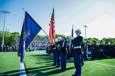 A military color guard standing in formation on a bright green turf football field during a graduation ceremony. Four individuals in blue uniforms, white helmets, and white gloves hold flags—including the American flag and the Pennsylvania state flag—and rifles. A large crowd sits in stadium bleachers labeled "NexTier Bank Stadium" in the background under a sunny sky.