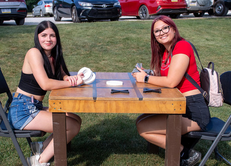 Two young women sitting at a wooden table on the grass, playing a large outdoor game of tic-tac-toe. They are smiling at the camera, with parked cars visible behind them.